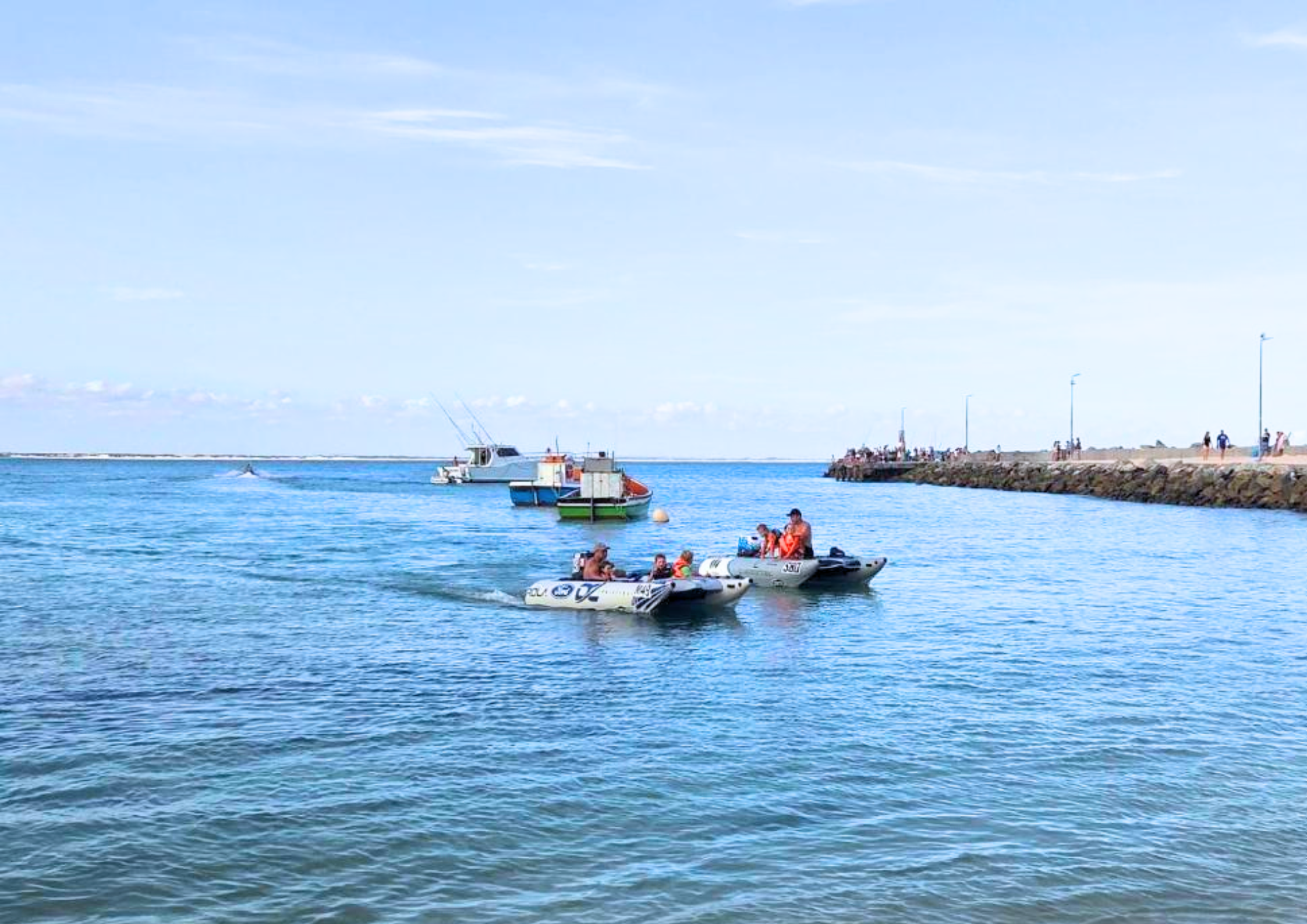 Two small inflatable boats with passengers on calm blue water near a harbour entrance, with fishing boats in the distance and people walking along a rocky breakwater.