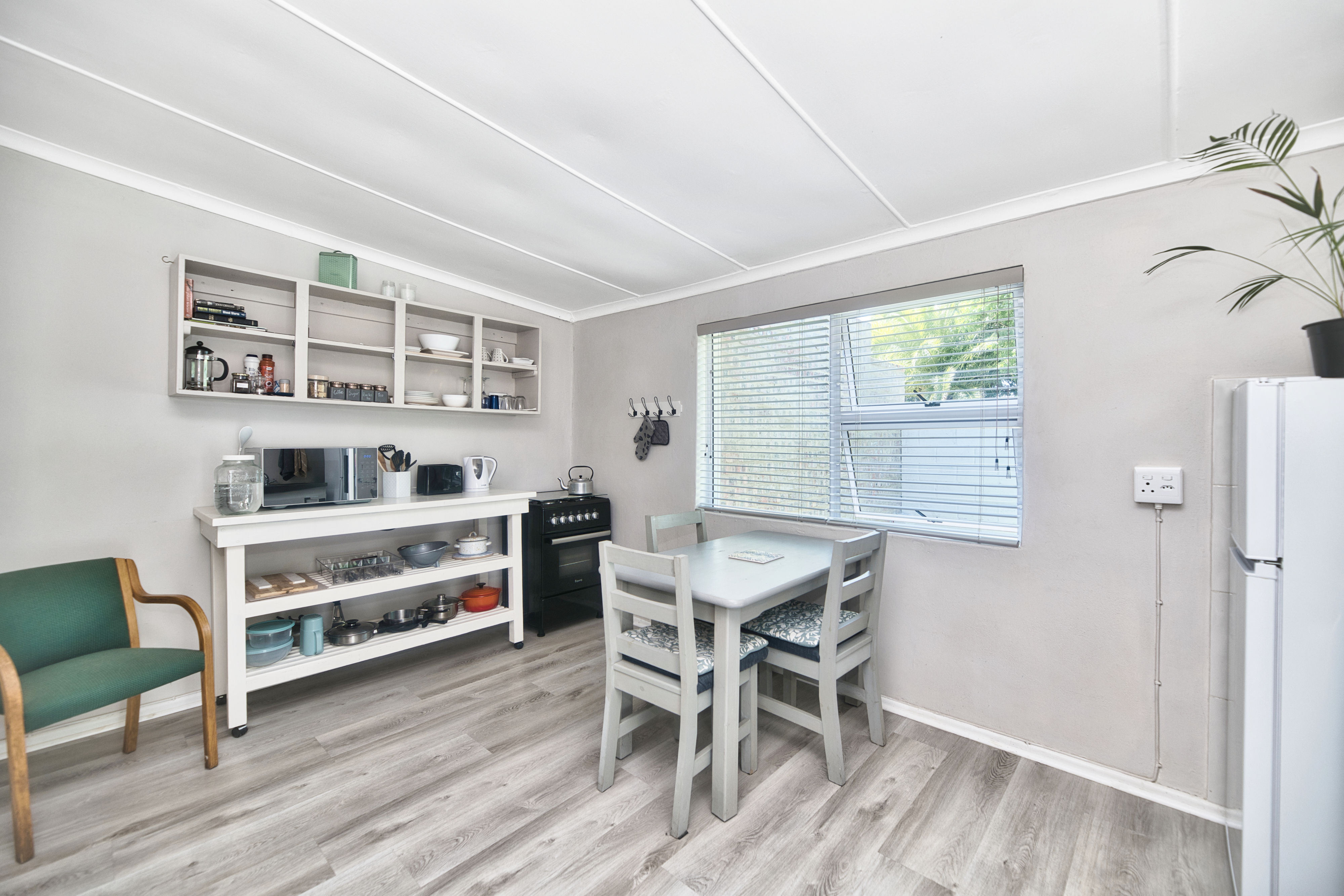 Bright kitchen with open shelving and cookware storage, a small dining table and chairs under a window with blinds, a freestanding stove, and a fridge next to a sink counter.