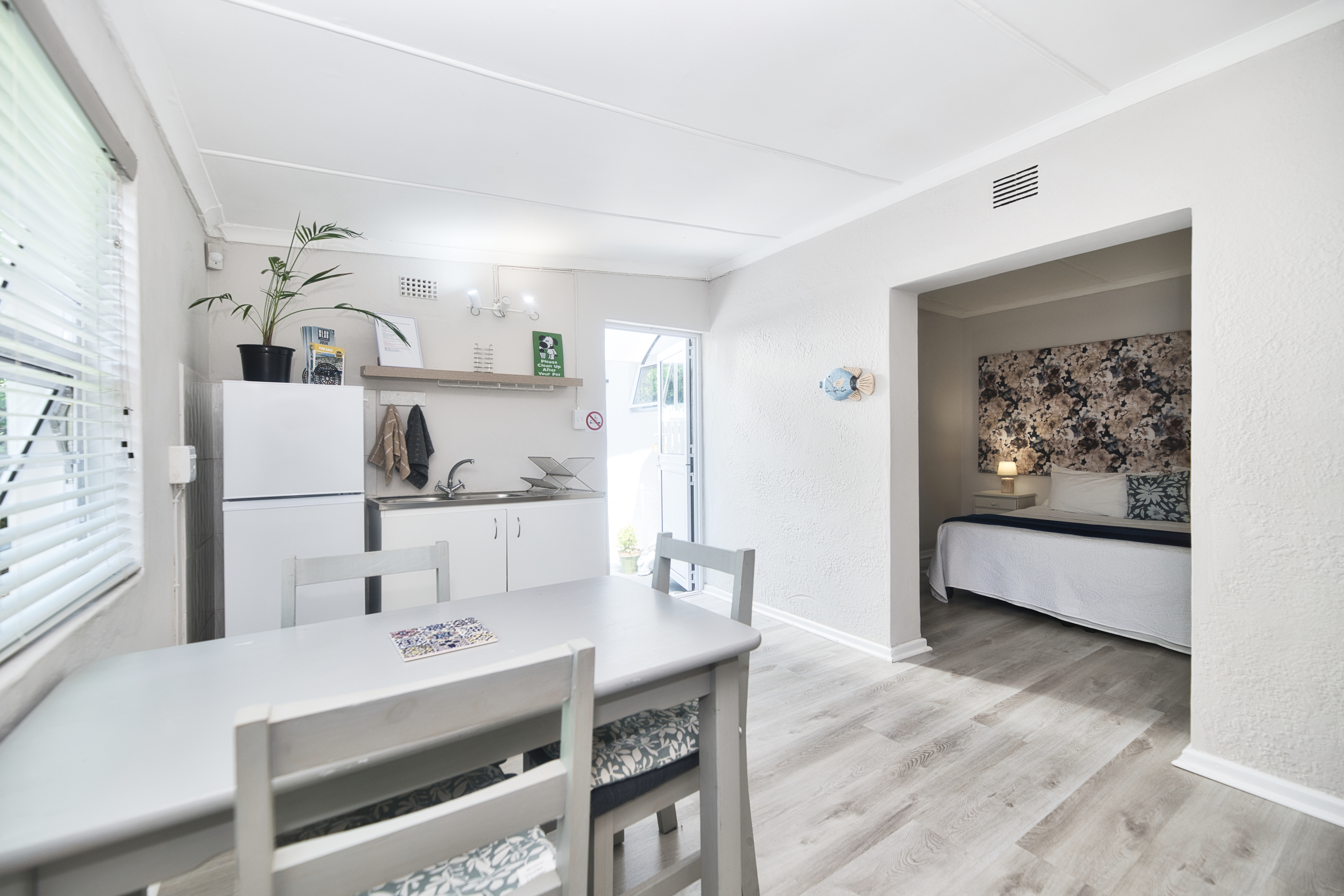 Small dining area with a table and chairs, a kitchenette with a fridge and sink, an open door to the outside, and an open doorway leading to a bedroom with a double bed.