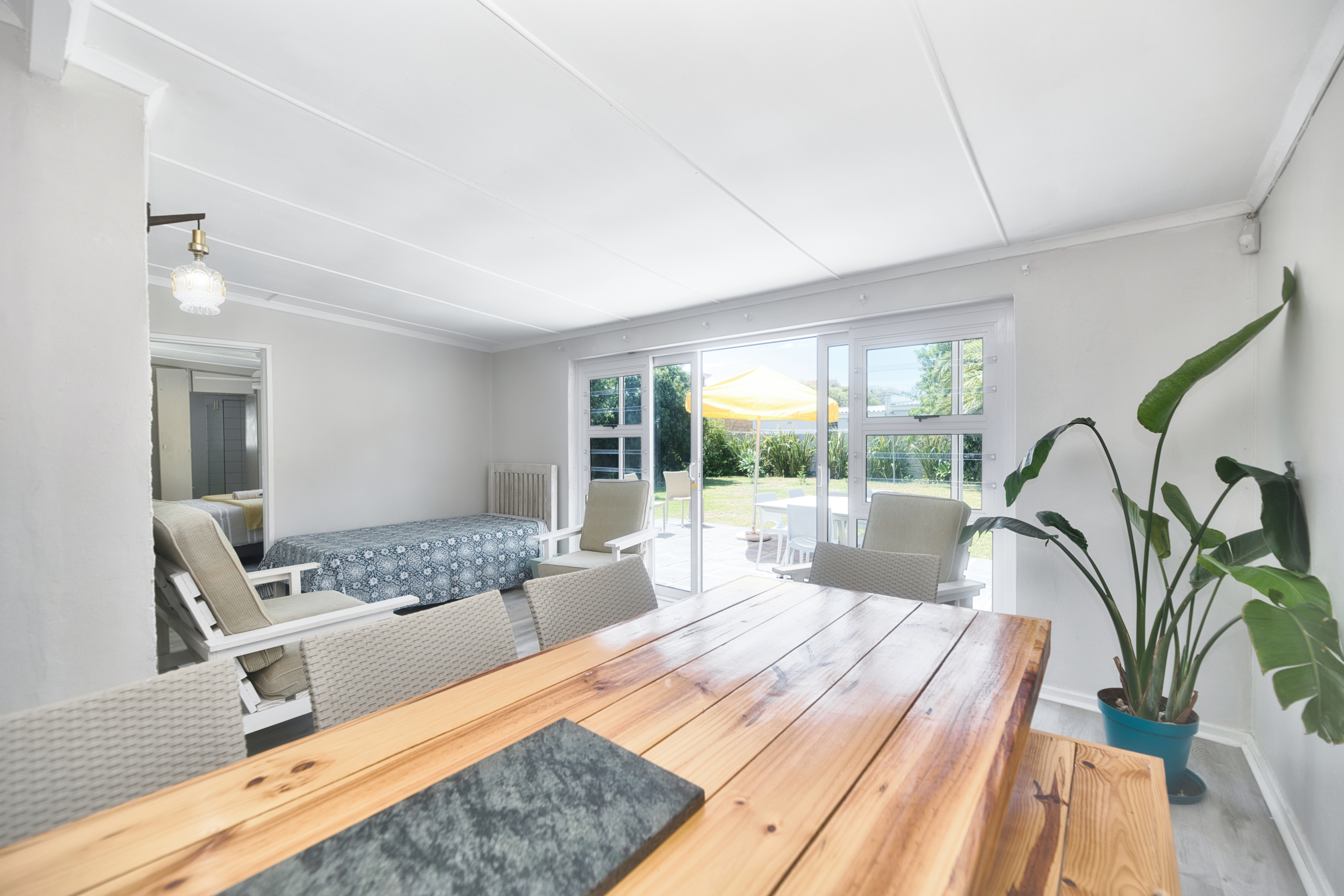 Bright dining area with a large wooden table and woven chairs, lounge seating to the left, a potted plant on the right, and sliding glass doors opening to a patio and garden.