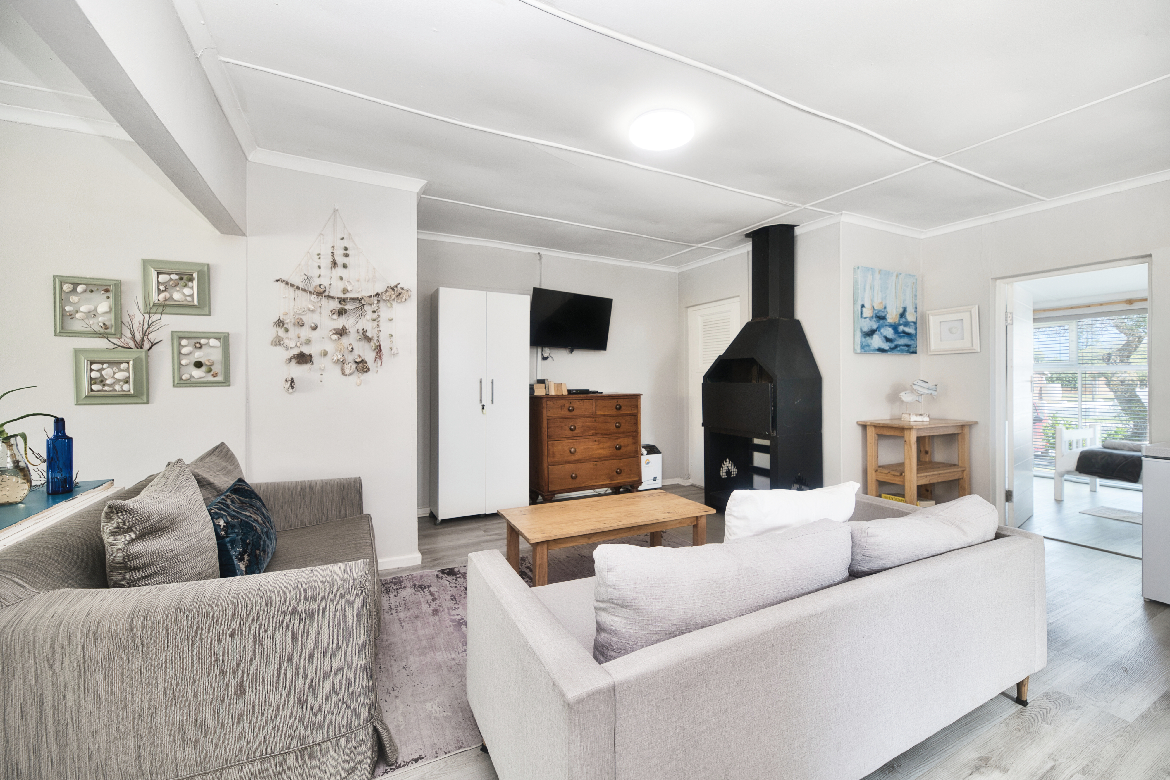 Living room with two sofas and a wooden coffee table, a wall-mounted TV, coastal wall art, and a large black indoor braai/fireplace in the corner.