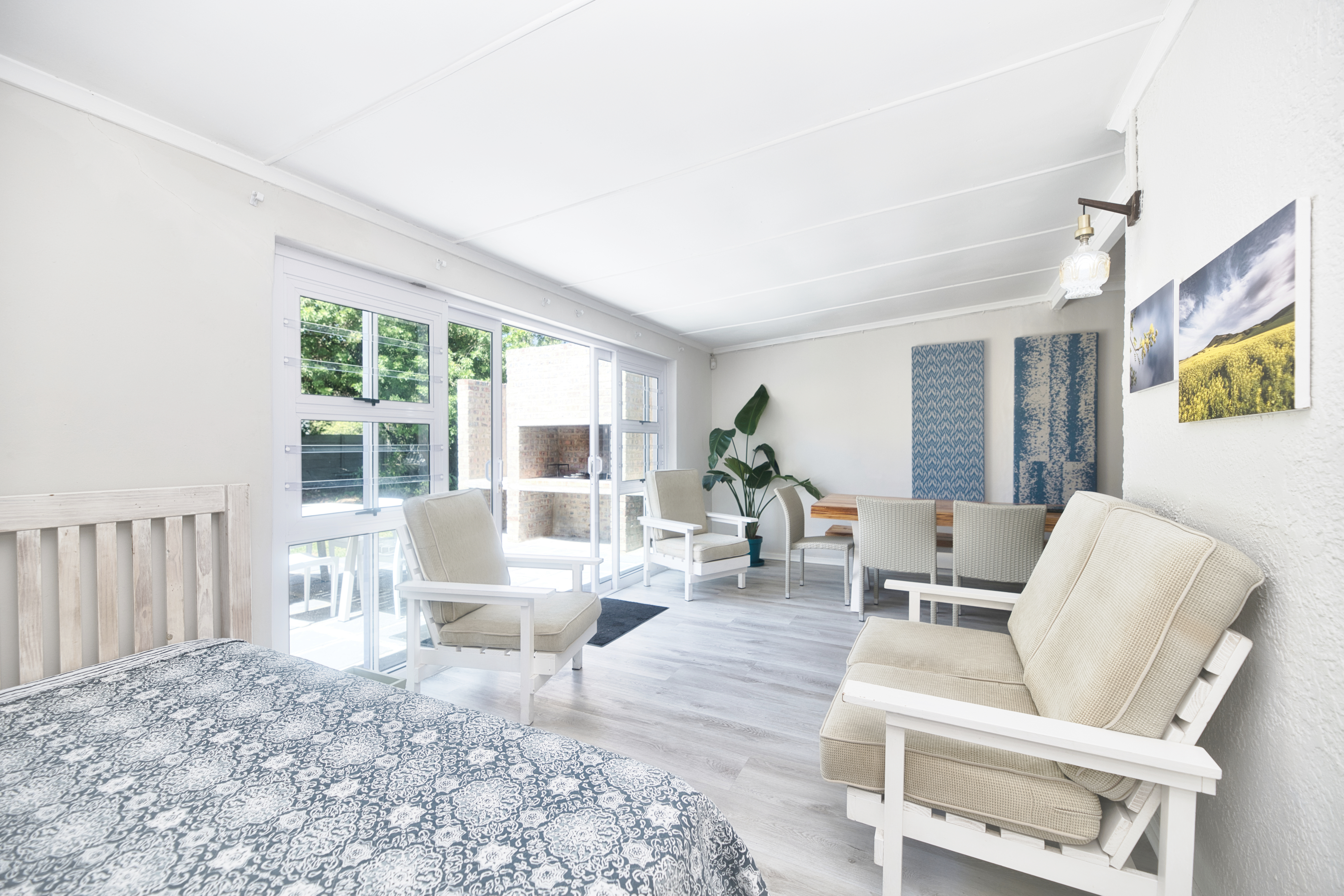 Bright open room with a bed on the left, cushioned lounge seating and chairs, a small dining table, and large sliding doors leading to an outdoor braai area.