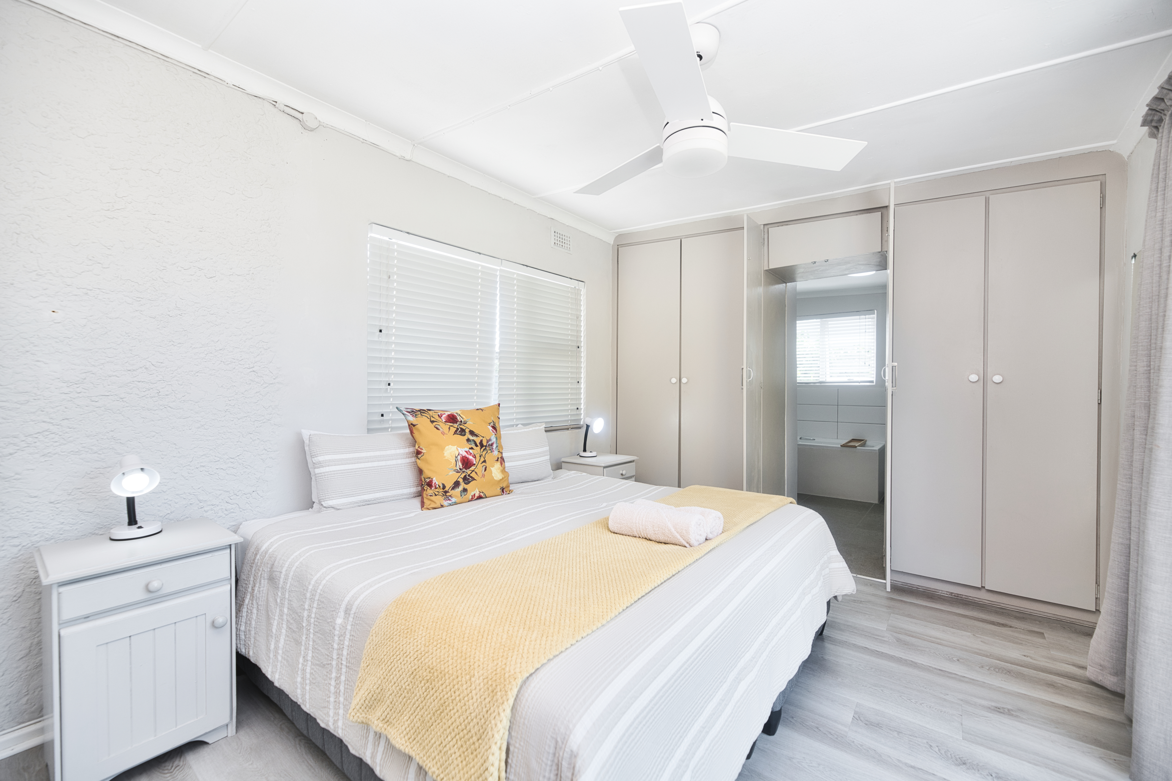 Double bedroom with a neatly made bed and yellow throw, bedside tables with small lamps, built-in cupboards, a ceiling fan, and an open doorway leading to an ensuite bathroom.