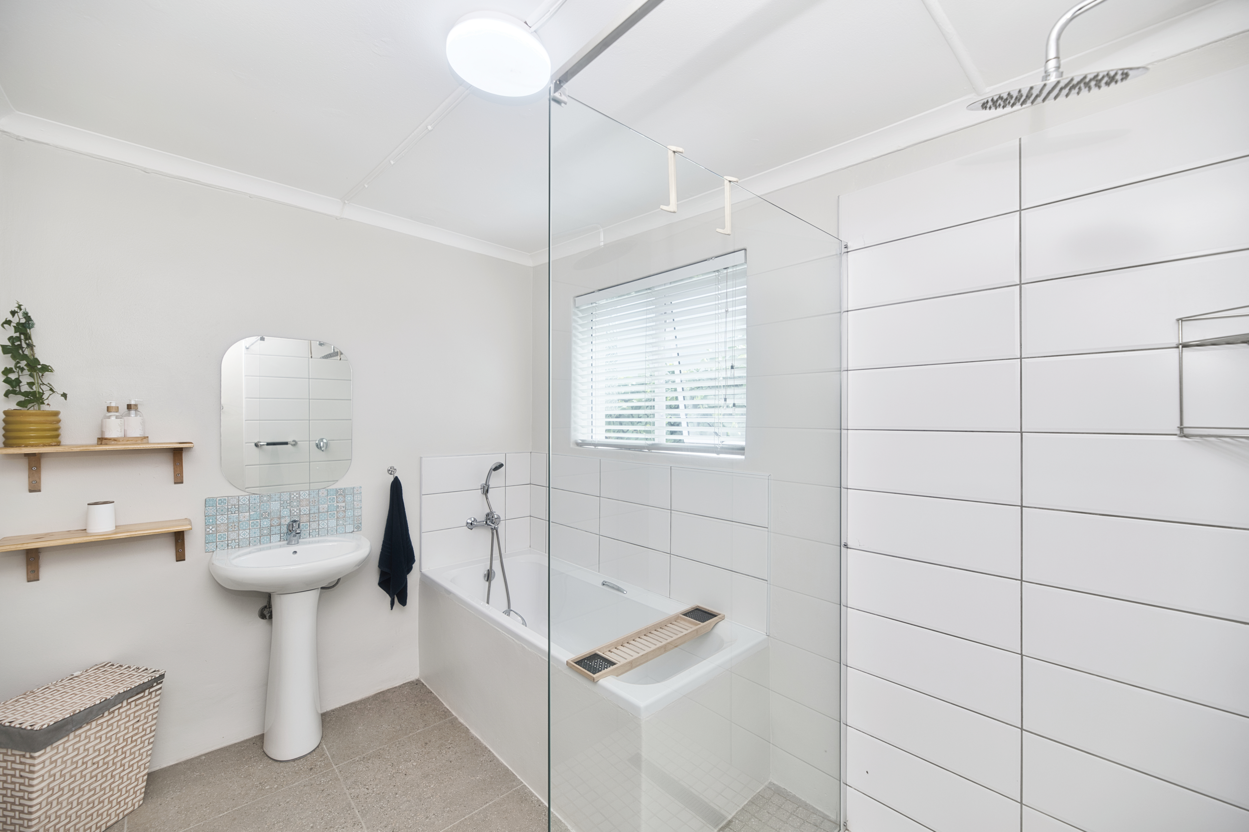 White bathroom with a bathtub and glass shower screen, a rain shower above, a pedestal sink with mirror, open shelves, and a window with blinds.