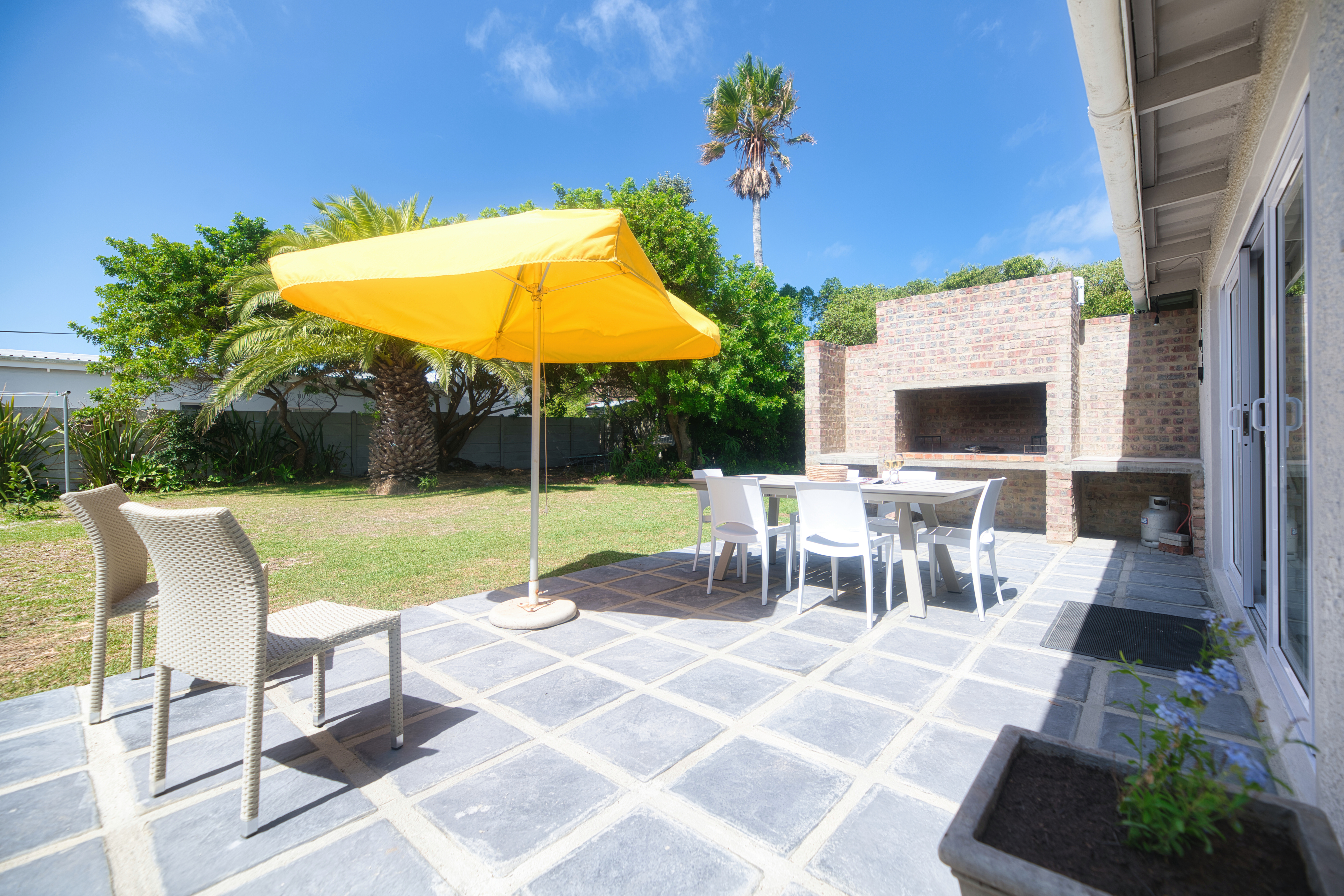Outdoor patio with a white dining table and chairs under a yellow umbrella, next to a brick built-in braai, with a grassy garden and trees in the background.