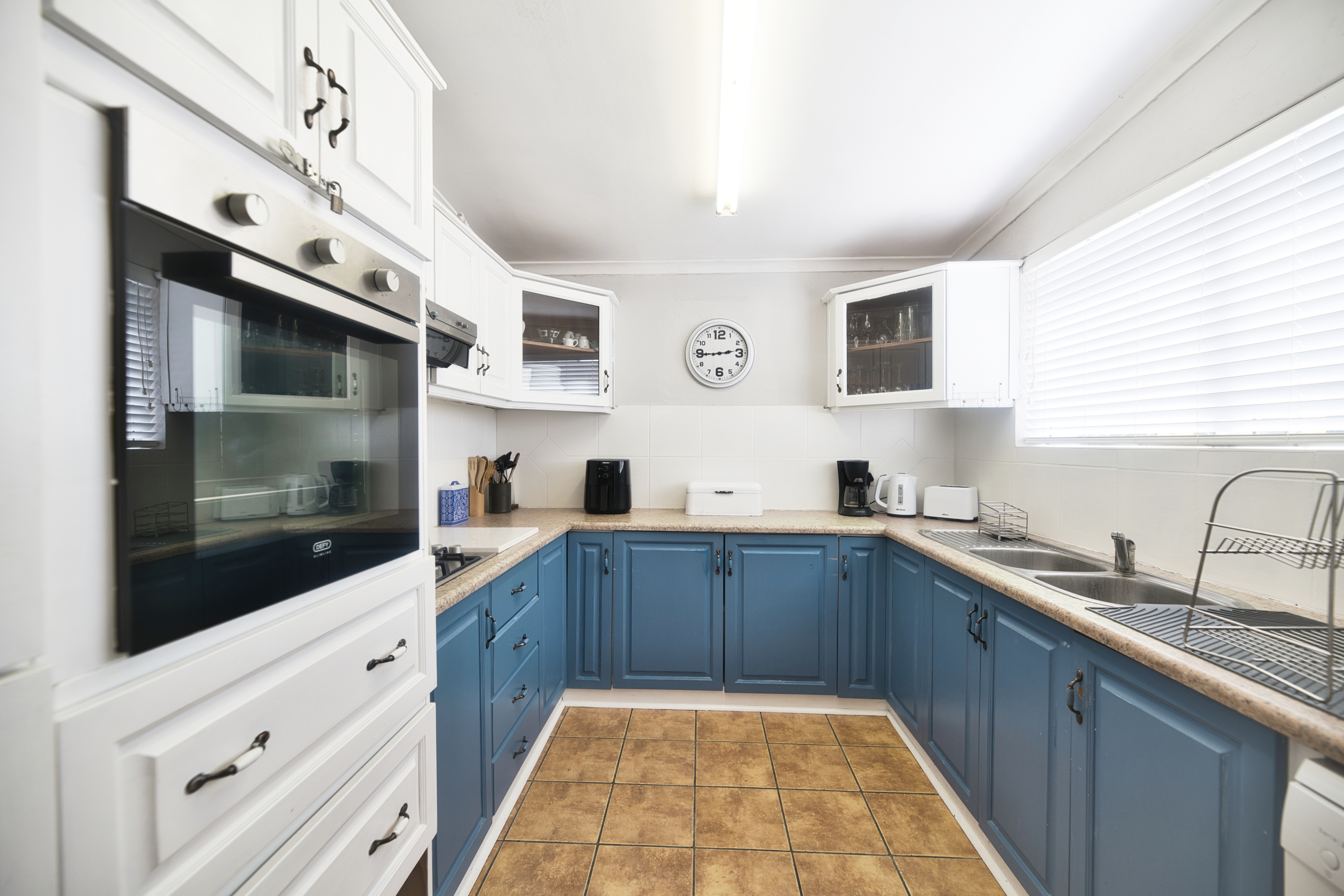 ompact U-shaped kitchen with white upper cupboards, blue lower cupboards, built-in oven, double sink under a window with blinds, and small appliances on the counter.