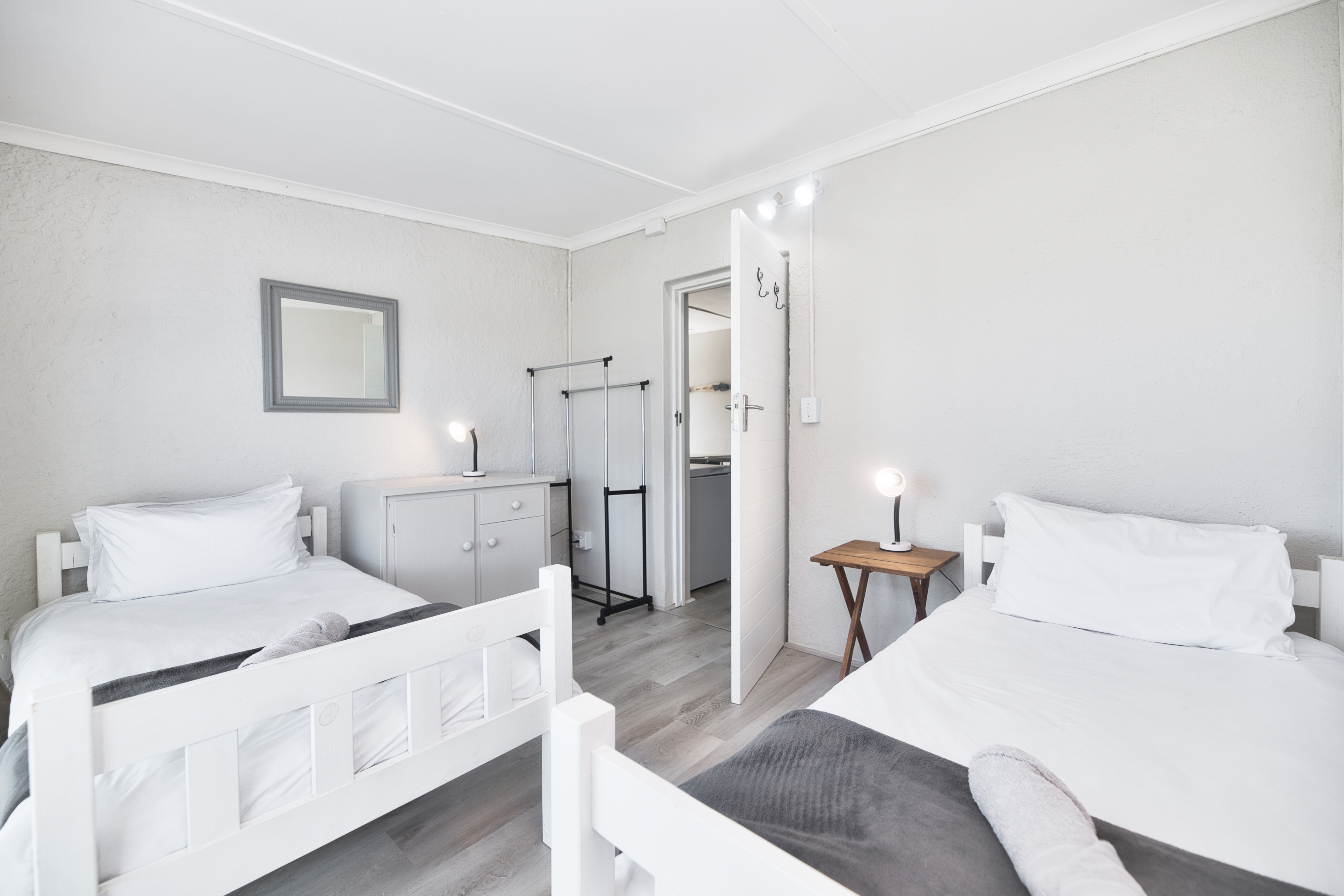 Simple twin bedroom with two single beds in white bedding, small bedside lamps, a compact cabinet, and an open door leading to another room.